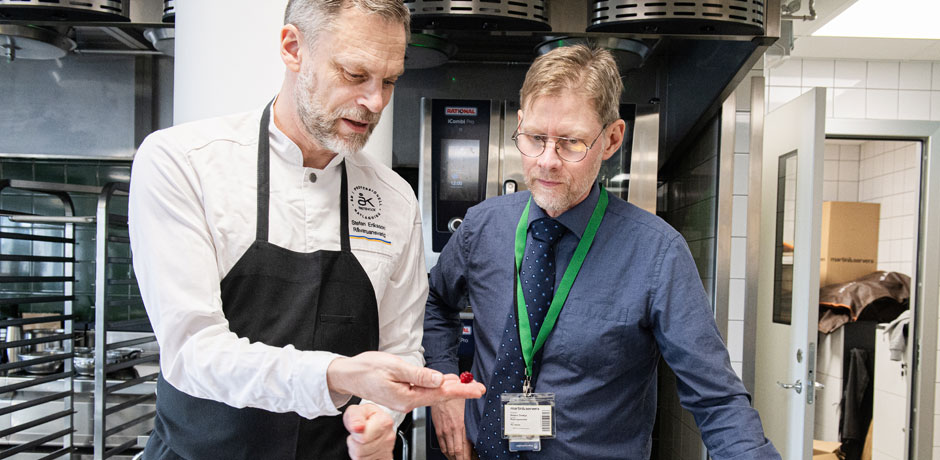 Två män, en som är kock och en som har skjorta och slips tittar och håller fram maten.