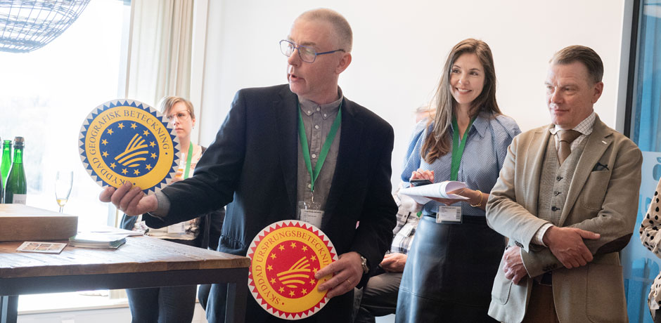 En man i kavaj håller upp logotyperna för geografisk beteckning. En kvinna och en man ser på.