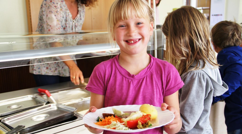 Flicka i sjuårsåldern som håller fram en tallrik med mat, i skolbespisningen. 