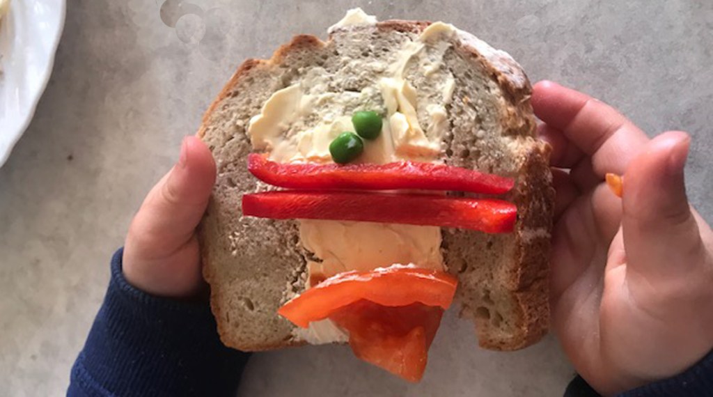 En smörgås med två ärtor, paprika och tomat format som ett ansikte. Smör i botten. 